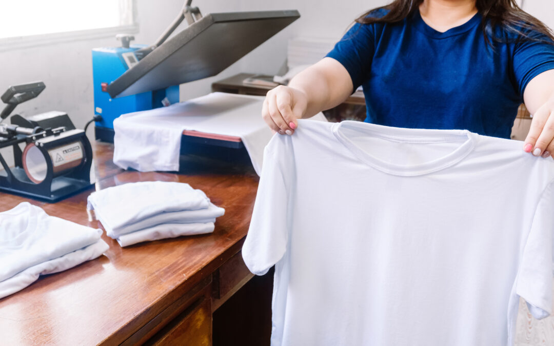 Female holding a white T shirt next to the cloths printing machine and T shirts on the wooden table