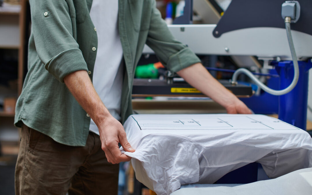 Cropped view of young craftsman holding t shirt with marking while working with screen printing machine in print studio at background, customer focused small business concept