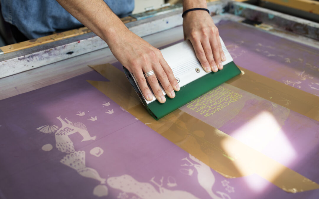 Close-up on a textile screen printer using a squeegee to print a pattern. The surface is purple and the design features white birds and circles, with a brown patch that has printed yellow text on it. The device is held by an adult whose gender cannot be determined and the hands are visible. This image would be ideal for projects about textile printing, crafts, small businesses, and art projects.