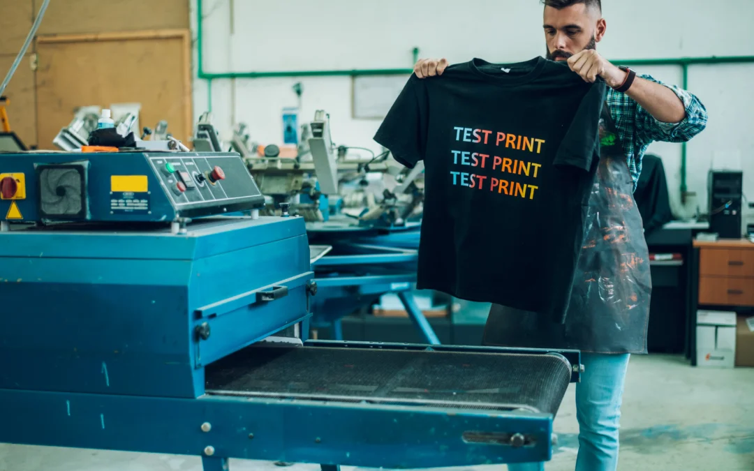 A man with dark hair and a beard is standing in a printing workshop, holding up a black t-shirt with the words "TEST PRINT" in colorful letters. He is wearing a blue and green plaid shirt with a dark apron. Behind him is a blue piece of printing equipment.