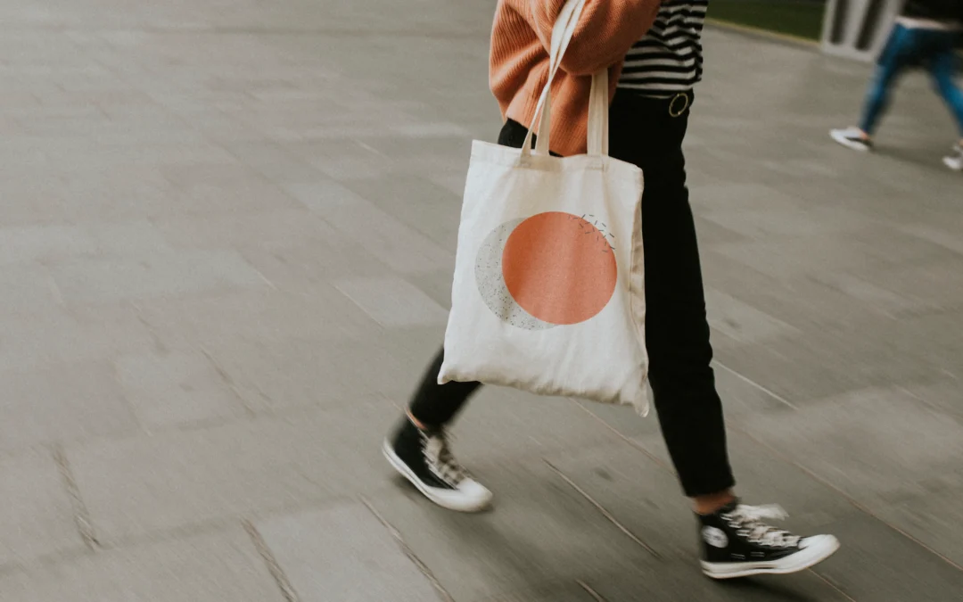 A stylish person walks along a city street carrying a tote bag. They are wearing dark pants, a horizontally striped shirt, an orange sweater, and high-top sneakers. The tote bag has a geometric design on it. The ground is grey and a blurry person walks behind.
