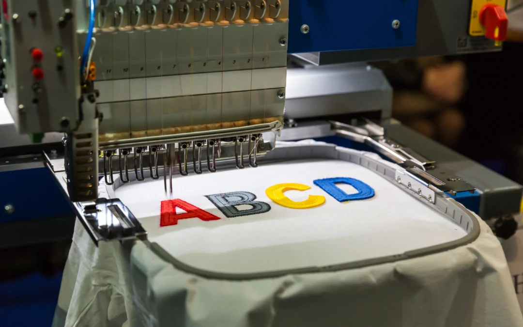 An embroidery machine is captured in a close-up shot, stitching colorful letters onto a piece of white fabric. The letters, A, B, C, and D, are in red, grey, yellow, and blue respectively. The machine is a complex piece of equipment with various metal components and settings. The photo conveys precision and craftsmanship.